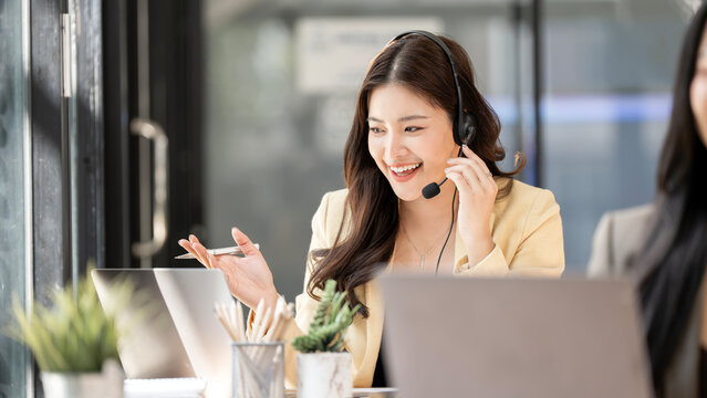 Professional Asian woman with headset working in call center, friendly customer service agent talking to client via video call, remote consultant in modern office.