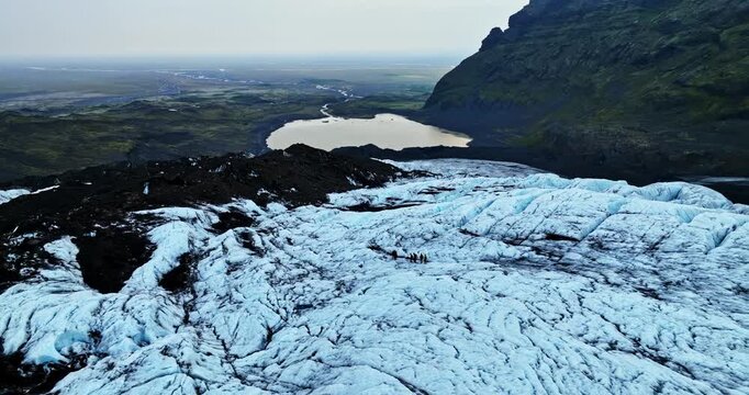 Drone view over Iceland icefield with tiny hikers crossing crevassed surface toward a milky meltwater lake beneath a steep mountain wall and dark volcanic ridges.