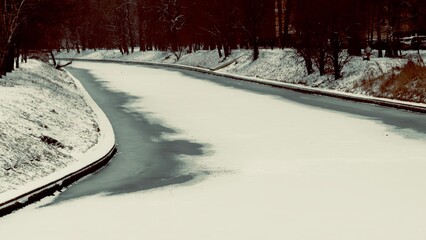 A serene winter scene featuring a snow-covered riverbank, with a winding river partially frozen, surrounded by bare trees and a tranquil atmosphere