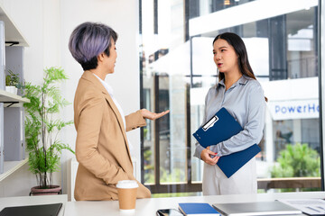 Confident mature business professional woman talking to younger female colleague at office table, speaking, gesturing, teaching, explaining work tasks. Elder mentor training intern.