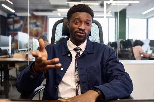 Portrait of young adult Black man sitting at desk gesturing with hand, looking into camera, office environment visible in background with colleagues working at computers