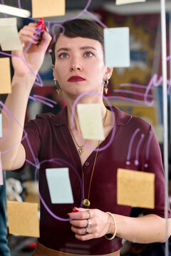 Caucasian young adult woman writing on transparent board with marker, surrounded by sticky notes, focusing on brainstorming or planning process in modern office environment