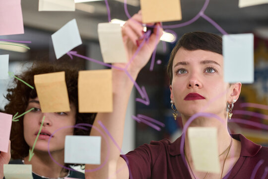 Young adult Caucasian women collaborating, placing sticky notes on glass wall, brainstorming ideas, focusing on project planning, teamwork in modern office