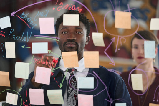 Young adult Black man brainstorming ideas on transparent board covered with colorful sticky notes and diagrams, young adult Caucasian woman standing in background observing process