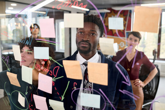 Young Black man standing in front of transparent board with sticky notes, collaborating with young Caucasian women and drawing diagrams during creative brainstorming session in modern office