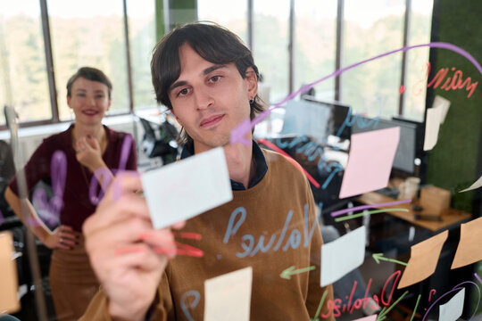 Caucasian young adult man writing on transparent board with colorful markers while Caucasian young adult woman standing in background observing brainstorming session in modern office
