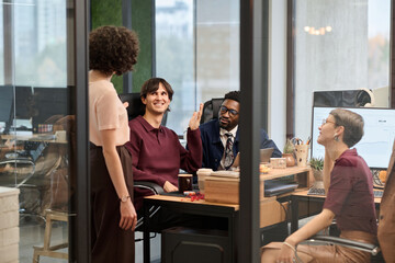 Diverse group of young adult men and women collaborating in modern office, Caucasian man gesturing while talking to standing Caucasian woman, Black man and Caucasian woman listening