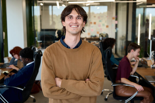 Portrait of young adult Caucasian man smiling with arms crossed in modern office, diverse group of coworkers collaborating in background, open workspace environment visible