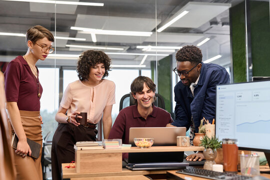 Diverse group of young adult men and women collaborating around laptop in modern office, smiling and discussing project while standing and sitting together, computer monitor visible in foreground