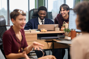 Group of young adult multiethnic colleagues collaborating around desk, Black man and Caucasian man smiling while working on laptop, two women gesturing and talking in foreground