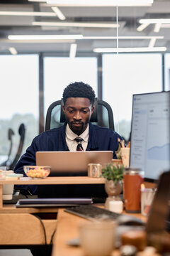 Young adult Black man sitting at desk working on laptop in modern office environment, focusing on computer screen with charts visible on monitor, surrounded by office supplies