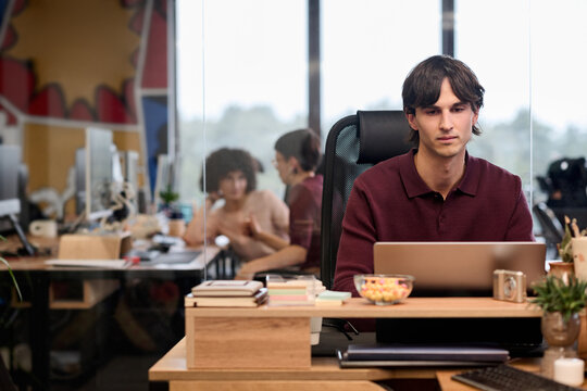 Young adult Caucasian man working on laptop at modern office desk, focusing on screen with colleagues collaborating in background, stack of books and potted plant on workspace