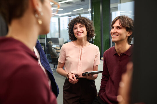 Young adult Caucasian woman holding digital tablet standing with young adult Caucasian man smiling and listening to colleague during informal meeting in modern office workspace