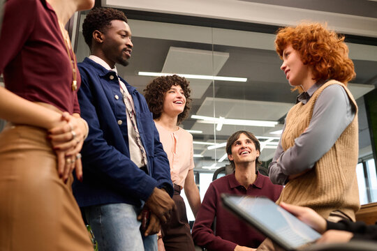 Diverse group of young adults standing and sitting in modern office, engaging in conversation, smiling and interacting with each other