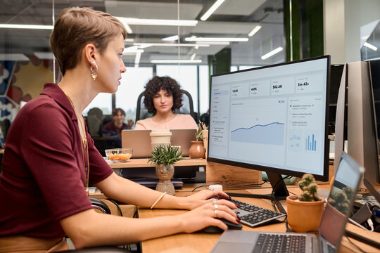 Caucasian young adult woman working at computer with business analytics dashboard on screen, multiethnic young adult colleagues visible in modern office workspace
