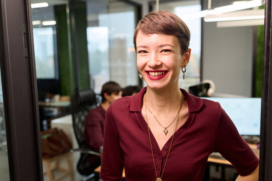 Portrait of young adult Caucasian woman smiling confidently while standing in modern office, showing short hair and jewelry, with colleagues working at computers in background