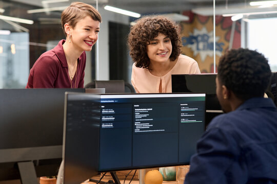Two young adult Caucasian women smiling and leaning over computer monitors engaging in conversation with young adult Black man sitting at workstation in modern office setting
