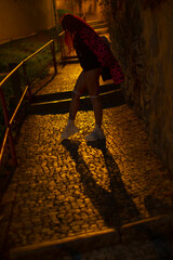 A stylish figure walks through a dimly lit urban path, casting intriguing shadows on cobblestones.