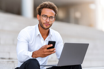 Businessman using smartphone and laptop outdoors, work and digital lifestyle concept.
