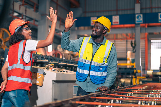 African factory worker man woman shake hand high five success. Industrial engineer partner male female wear safety helmet vest in manufacturing plant. Heavy metal machine background. Happy diversity