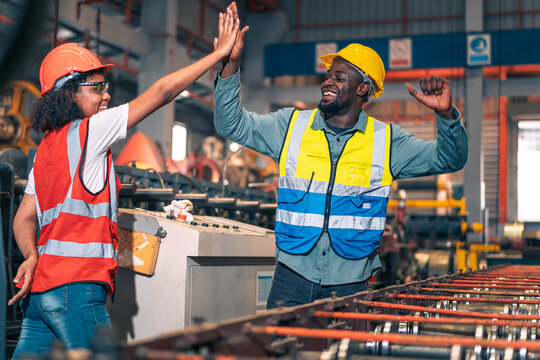 African factory worker man woman shake hand high five success. Industrial engineer partner male female wear safety helmet vest in manufacturing plant. Heavy metal machine background. Happy diversity
