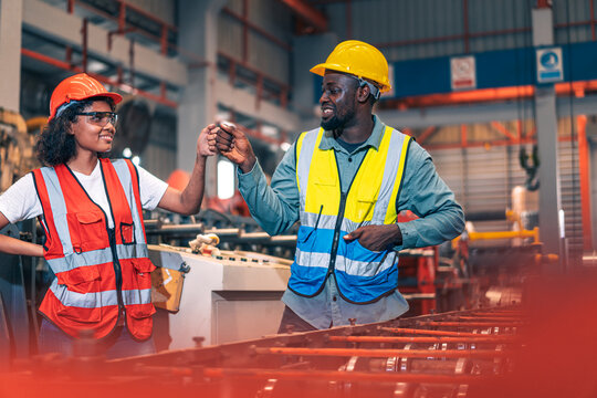 African factory worker man woman fist bump team success. Industrial engineer partner male female knuckle bump power in manufacturing plant. Safety helmet vest heavy metal machine background. Diversity