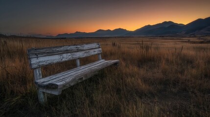 Fototapeta premium An old wooden bench sits on dry grassy terrain during sunrise or sunset with warm hues in the sky and distant mountain s