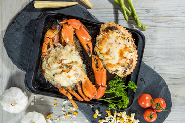 A black tray with two crabs baked in creamy cheese topping surrounded by fresh vegetables and garlic cloves.