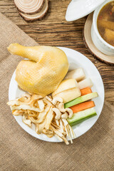 A plate of boiled chicken served with roots, berries, and herbs beside a ceramic soup pot.
