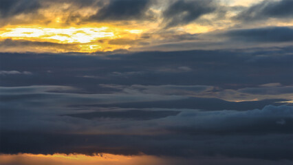Fototapeta premium Moody Layered Evening Sky With Bands Of Dark Blue Gray Clouds Parting To Reveal Strips Of Golden Light Near The Horizon, Creating A Dramatic, Reflective And Atmospheric Sunset Scene
