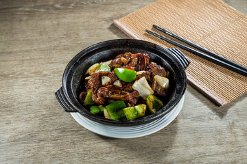 A clay pot filled with braised beef chunks cooked with green peppers and onions served on a wooden mat.