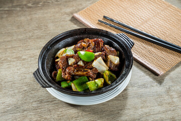 A clay pot filled with braised beef chunks, green peppers, and onions in rich sauce, served on a wooden table with chopsticks.