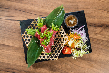 A black plate with raw beef slices wrapped around greens, served with salad and dipping sauce on a wooden table for a stylish presentation.