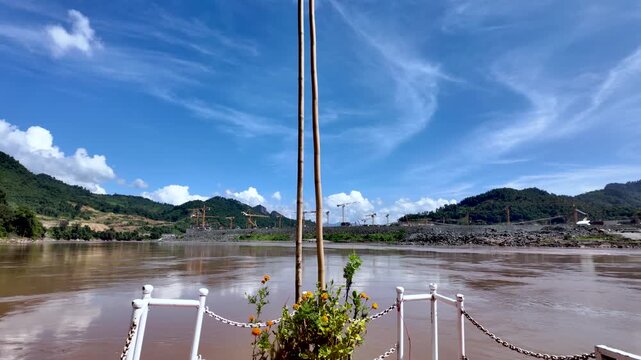 Bow of a traditional boat on the Mekong River, passing a large dam construction site in Laos
