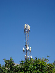 A tall cellular tower stands against a vivid blue sky, partially obscured by lush green foliage, showcasing modern communications technology.