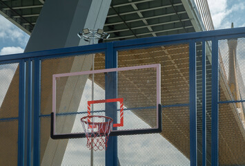 A blue steel grid fence frames a fiberglass basketball backboard with an orange rim and net, set against the backdrop of the industrial Ring Road Bridge. Copy space.