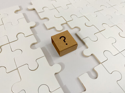 A small cardboard cube with a printed question mark sits on a white jigsaw puzzle, representing missing information, business uncertainty, and finding strategic solutions.