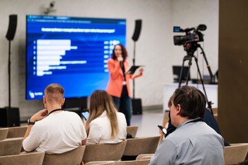 Presenter speaks to seated attendees while large screen shows charts, camera records session, attendees watch, take notes, and use phones during business conference.