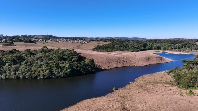 High-altitude aerial side reveal of a serene water reservoir surrounded by golden winter vegetation. Features a distant view of Sao Francisco de Paula in Serra Gaucha, Brazil.