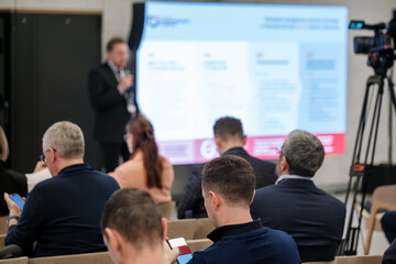 Audience attends a conference as a speaker presents on a large screen. Camera crew captures the event in a modern venue.