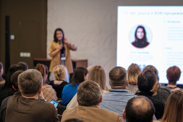 Presenter speaks to crowd during conference session. Audience seated listening while large screen shows profile slide. Collaborative learning and professional exchange emphasized.