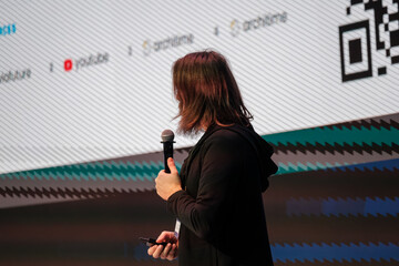 Speaker on stage delivers talk with microphone in hand while standing before large screen with diagonal pattern and QR code design. Confident professional moment during conference presentation.