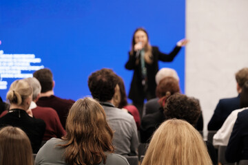 Speaker delivers presentation to attentive crowd in conference room. Blue backdrop creates modern setting. Audience listens closely feels curious inspired focused during talk.