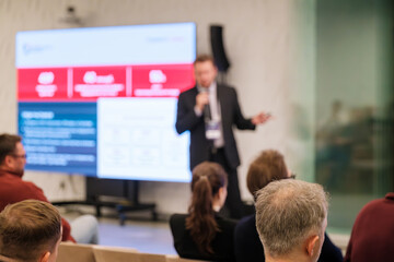 Blurry photo shows presenter in suit delivering speech to seated crowd during conference with large screen slides.
