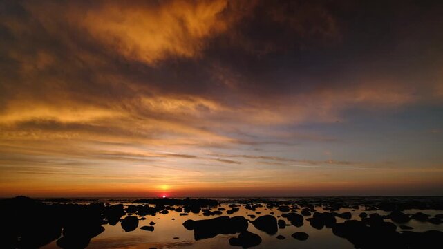 Peaceful time-lapse style footage of sun slowly setting behind sea horizon, coloring dramatic clouds in orange and gold and reflecting in calm water with dark rocks in foreground
