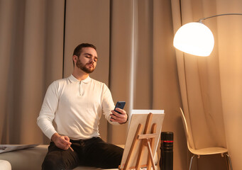 A young male artist photographs a painting on canvas with his smartphone while sitting in a cozy studio workshop with soft lighting.