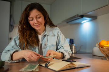 Woman managing household finances, counting cash money and planning budget, allocating funds for bills and savings, sitting at kitchen counter with notebook
