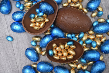 Selection of colourful foil wrapped chocolate easter eggs in blue, and gold. Large halves of a brown milk chocolate egg have mini eggs inside, on a grey wooden background.