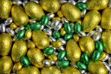 Large & small yellow, gold, green and silver spring colours of foil wrapped chocolate easter eggs, against a black background.
