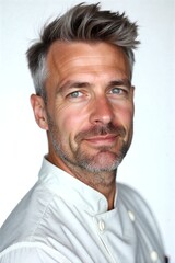 Confident chef smiles warmly, gray hair styled neatly, white coat and black apron against clean white background.